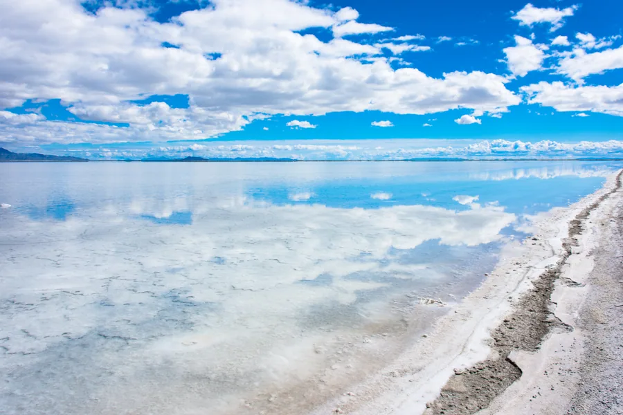 The cracked edge of the salt crust where it meets standing water, with the mountains reflected beyond.