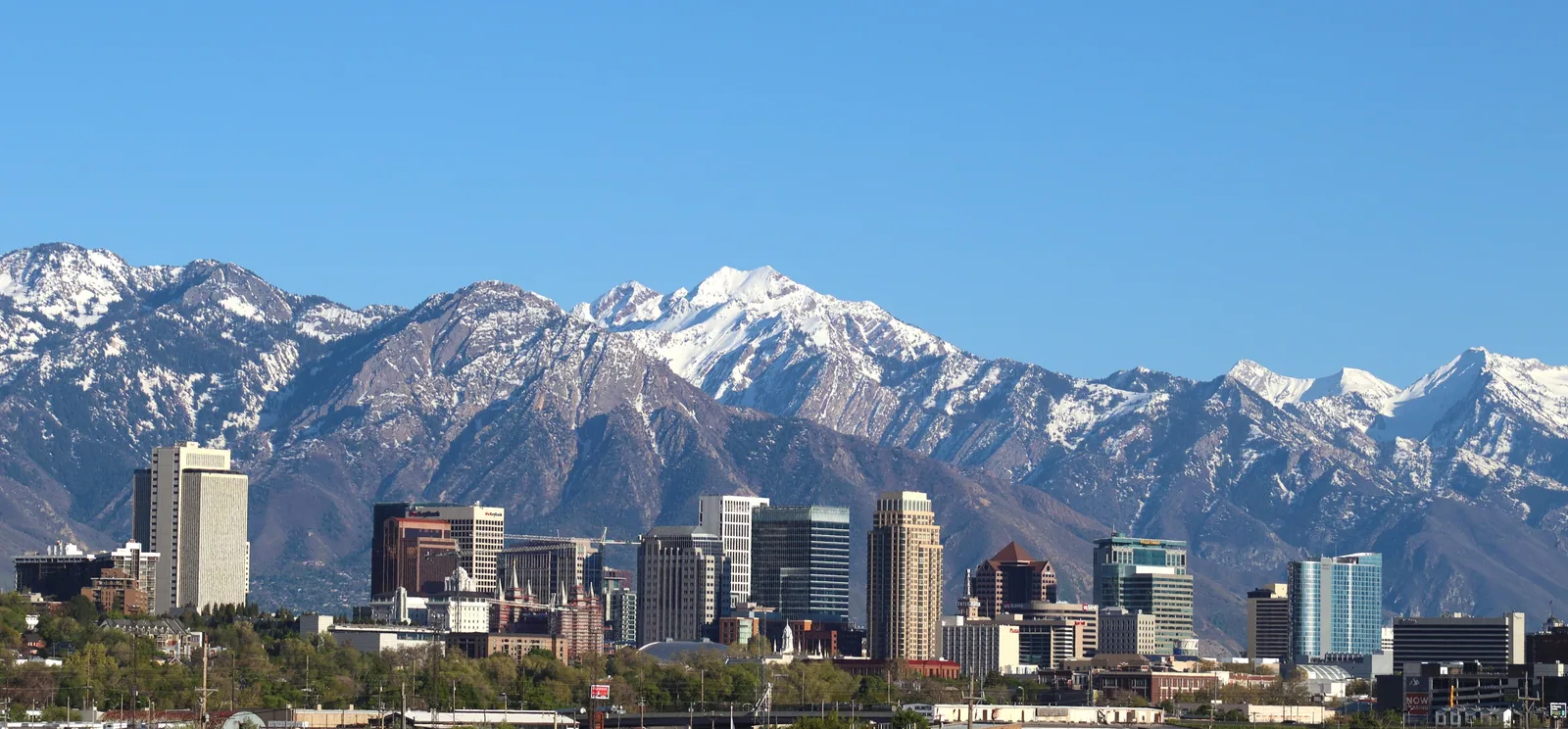 Downtown Salt Lake City on a clear spring day, the snow-covered peaks of the Wasatch Range rising directly behind the towers.